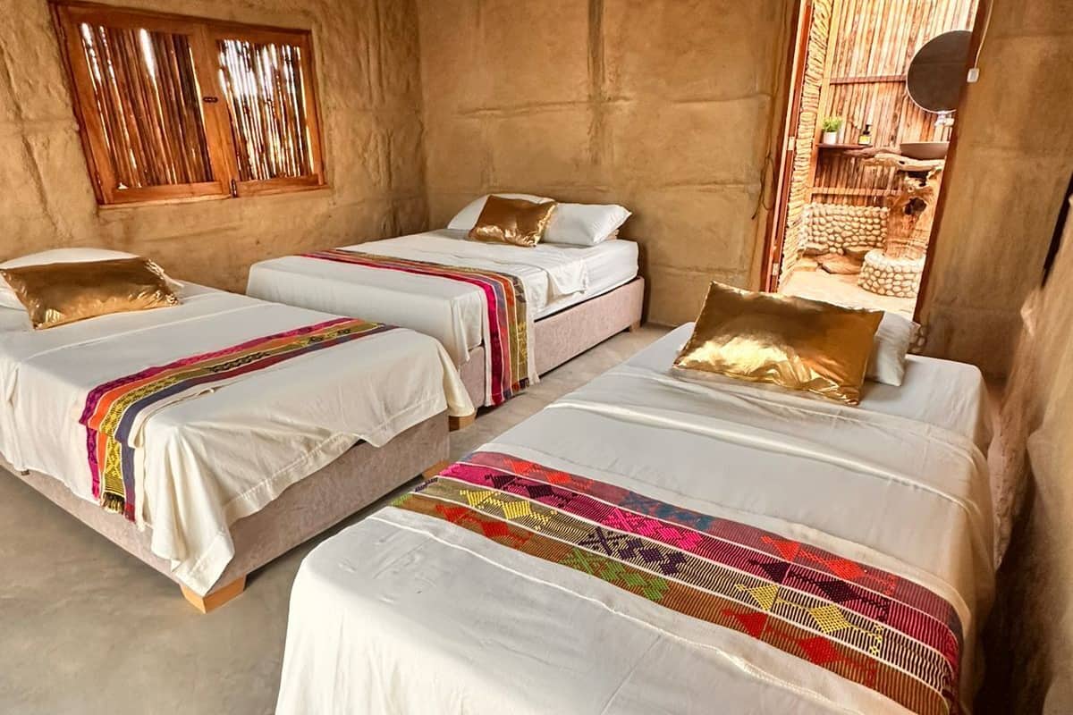Une chambre avec trois lits, chacun recouvert de draps blancs et de couvertures colorées. On y trouve un luminaire suspendu en osier et une fenêtre avec des stores en bois.