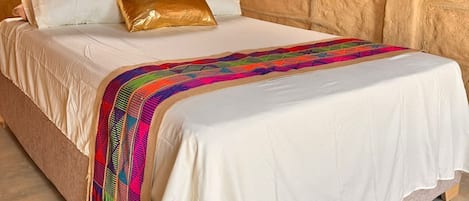 A bed with white linens, a colorful blanket, and a gold pillow in a room with stone walls and a corrugated metal ceiling.