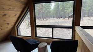 Een chalet met twee zwarte stoelen, een houten stronk en uitzicht op een bos door grote ramen.