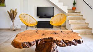 Un salon moderne avec une table basse en bois au design original, deux fauteuils jaunes, un téléviseur à écran plat et un escalier.