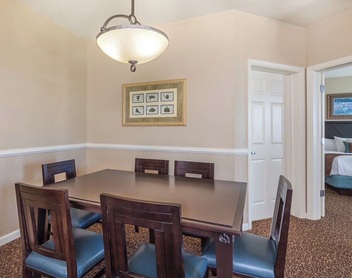 Un comedor con una mesa rectangular y seis sillas, una araña y un cuadro en la pared.