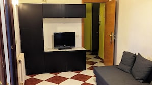 En kompakt opholdsstue med et skakternet gulv, en sofa, et fjernsyn på et sort skab og en radiator.