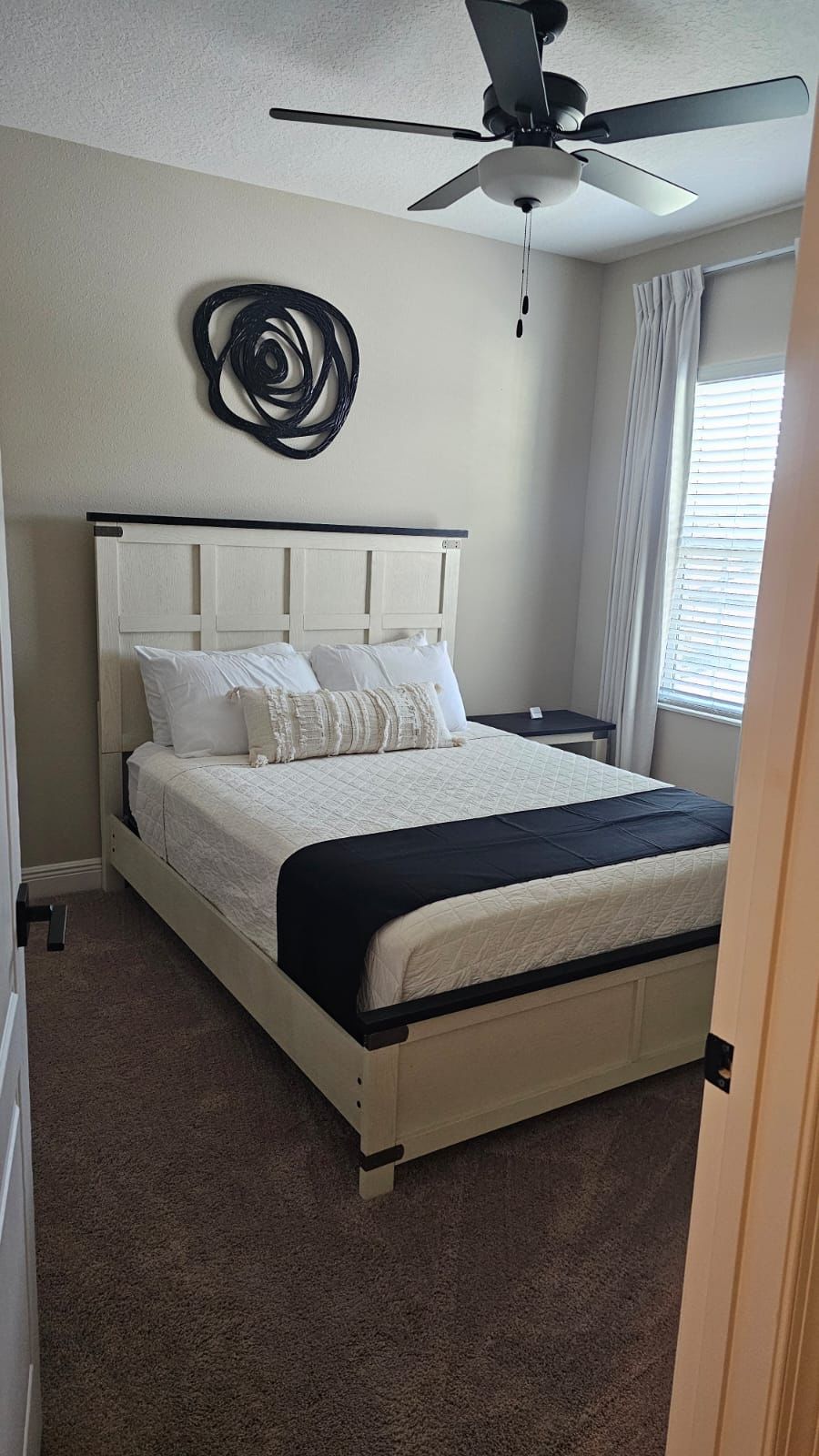 A bedroom with a bed, a ceiling fan, a wall decoration, a nightstand, and a window with curtains.