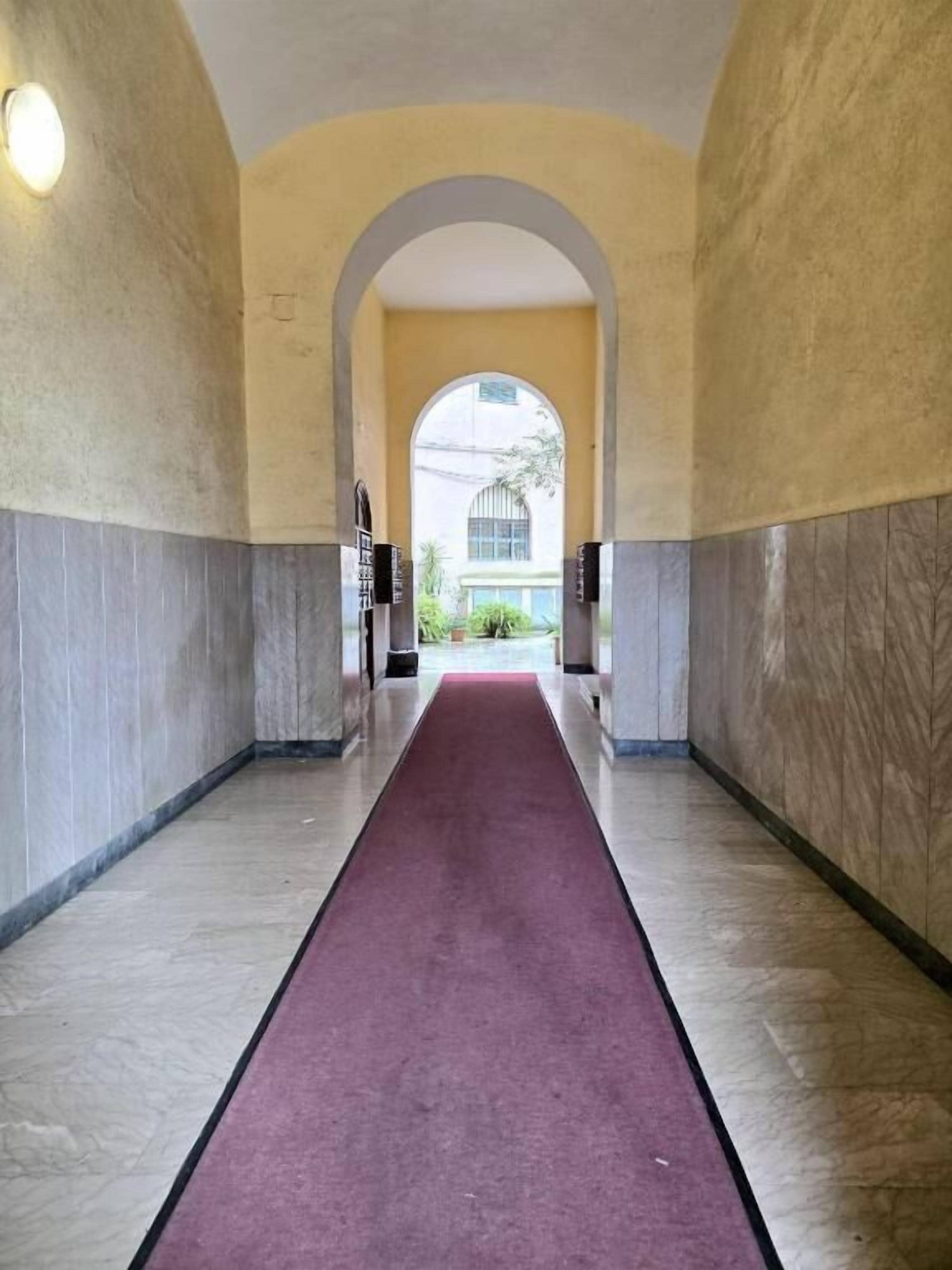 A hallway with a marble floor, a red carpet, and an arched doorway leading to a building.