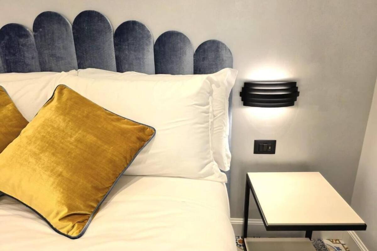A bed with white linens and a mustard-colored pillow, a wall-mounted light fixture, and a small bedside table.