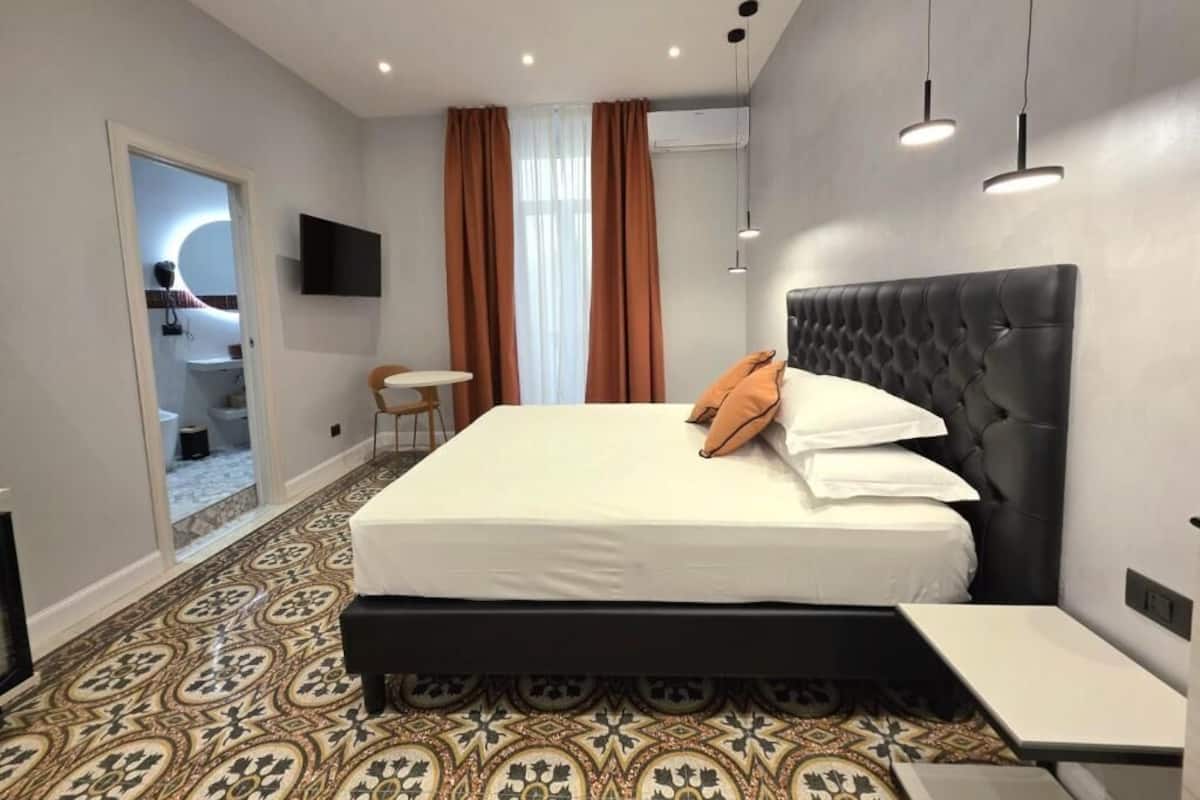 A modern hotel room with a large bed, a small bedside table, a TV mounted on the wall, and a patterned carpet.