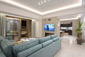 Ein modernes Wohnzimmer mit grauem Sofa, einem Flachbildfernseher und einem Essbereich, wobei im Hintergrund eine Küche zu sehen ist.