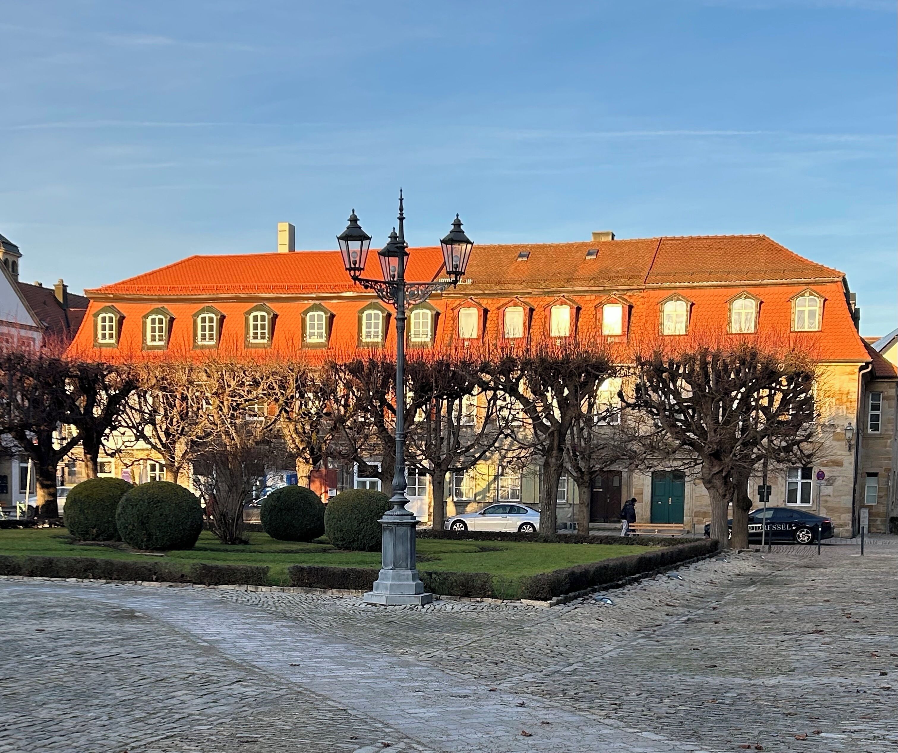 Ein historisches Gebäude mit rotem Dach und zahlreichen Fenstern, umgeben von Bäumen und einer Kopfsteinpflasterstraße.