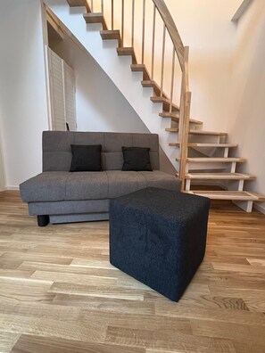Een moderne woonkamer met een grijze bank, zwarte kussens, een houten trap en een houten vloer.