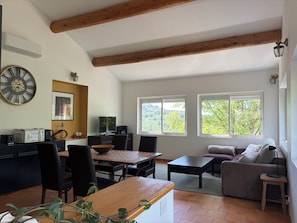 Ein großzügiger Wohnbereich mit Esstisch, Stühlen, einer Couch und einem Fernseher. Fenster bieten Blick auf Bäume.