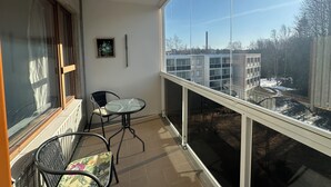 A balcony with a view of buildings and trees, a chair, a table, and a framed picture on the wall. - Spacious 2 Bedroom 75 sqm Kannelmäki (Helsinki)