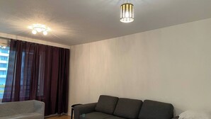 A modern living room with a grey sofa, a small white chair, and a large window with dark curtains. - Lovely 2-br 77m2 Apart Pasila-train Station (Helsinki)