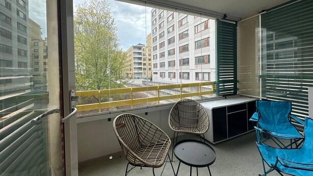 A balcony with a view of buildings and trees, featuring a desk, two wicker chairs, and a blue chair. - Lovely 2-br 77m2 Apart Pasila-train Station (Helsinki)
