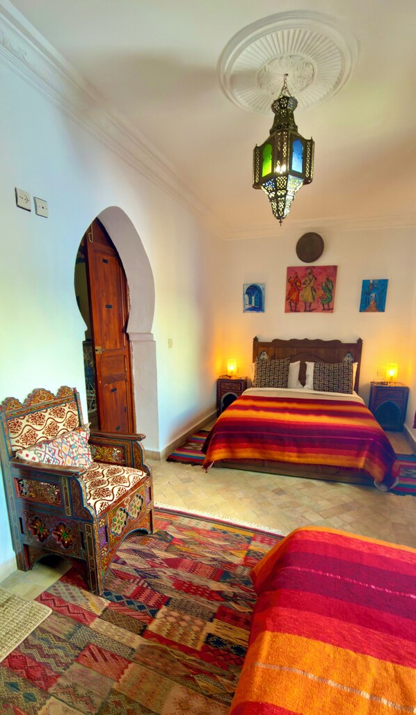 A bedroom with a bed, a chair, a carpet, a chandelier, and a wall with artwork.