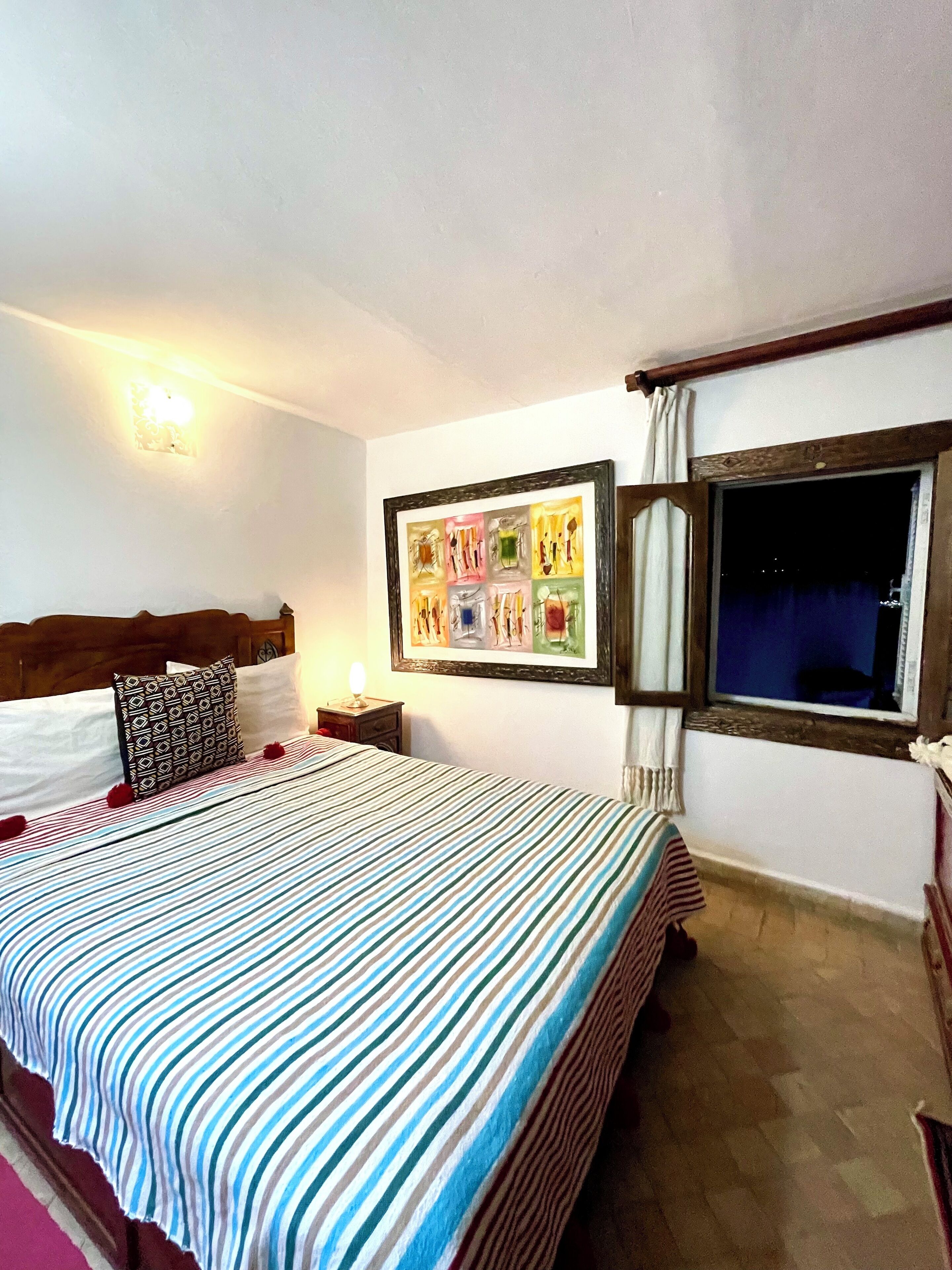 A hotel room with a large bed, a nightstand, a framed artwork on the wall, and a window with curtains.