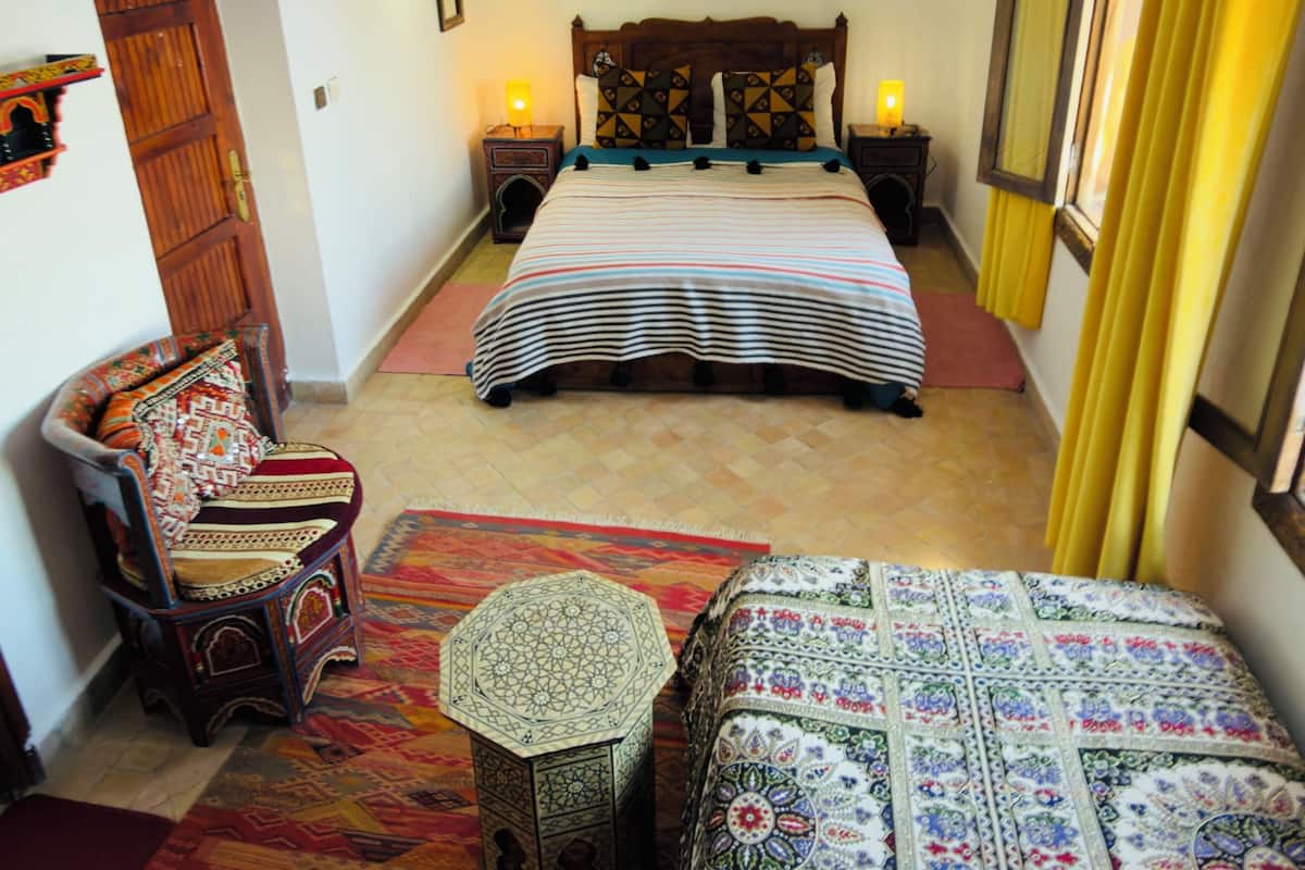 Quarto com cama, mesas de cabeceira, uma cadeira, uma pequena mesa e um tapete estampado.