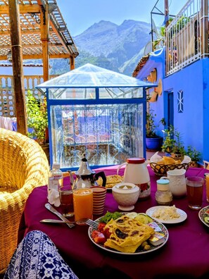 A table set with food and drinks, a wicker chair, and a view of mountains. - Dar Sababa (Chefchaouen)