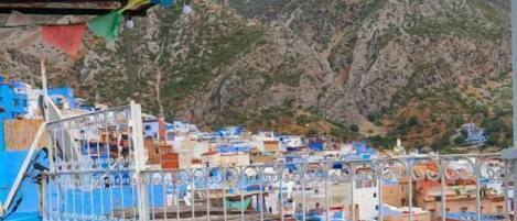 Un balcón con mobiliario de mimbre, vista a un pueblo de edificios azules y montañas al fondo.