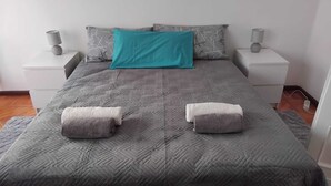 A neatly made bed with a grey quilt and two towels, a teal pillow, and two bedside lamps on white nightstands. - "Valeri" Vacation Rental in Sondrio - Peaceful Stay Near City Center (Sondrio)