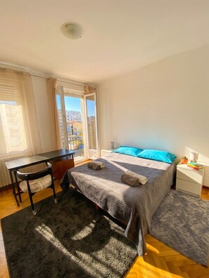 Ein Schlafzimmer mit Bett, Schreibtisch, Stuhl, kleinem Tisch und einem Fenster mit Blick auf Gebäude.