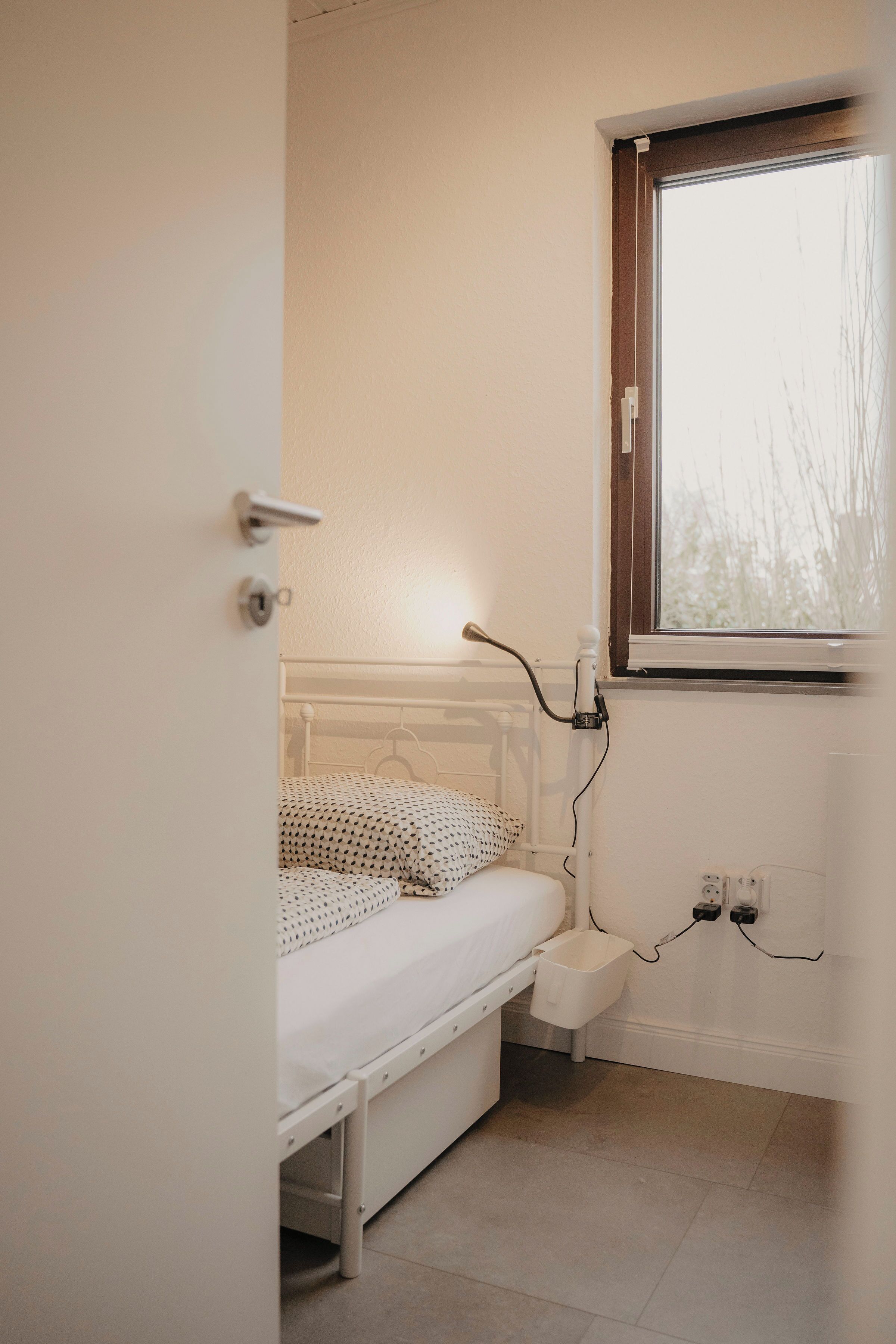 Une petite chambre avec un lit simple, une fenêtre, une table de chevet et une prise électrique.
