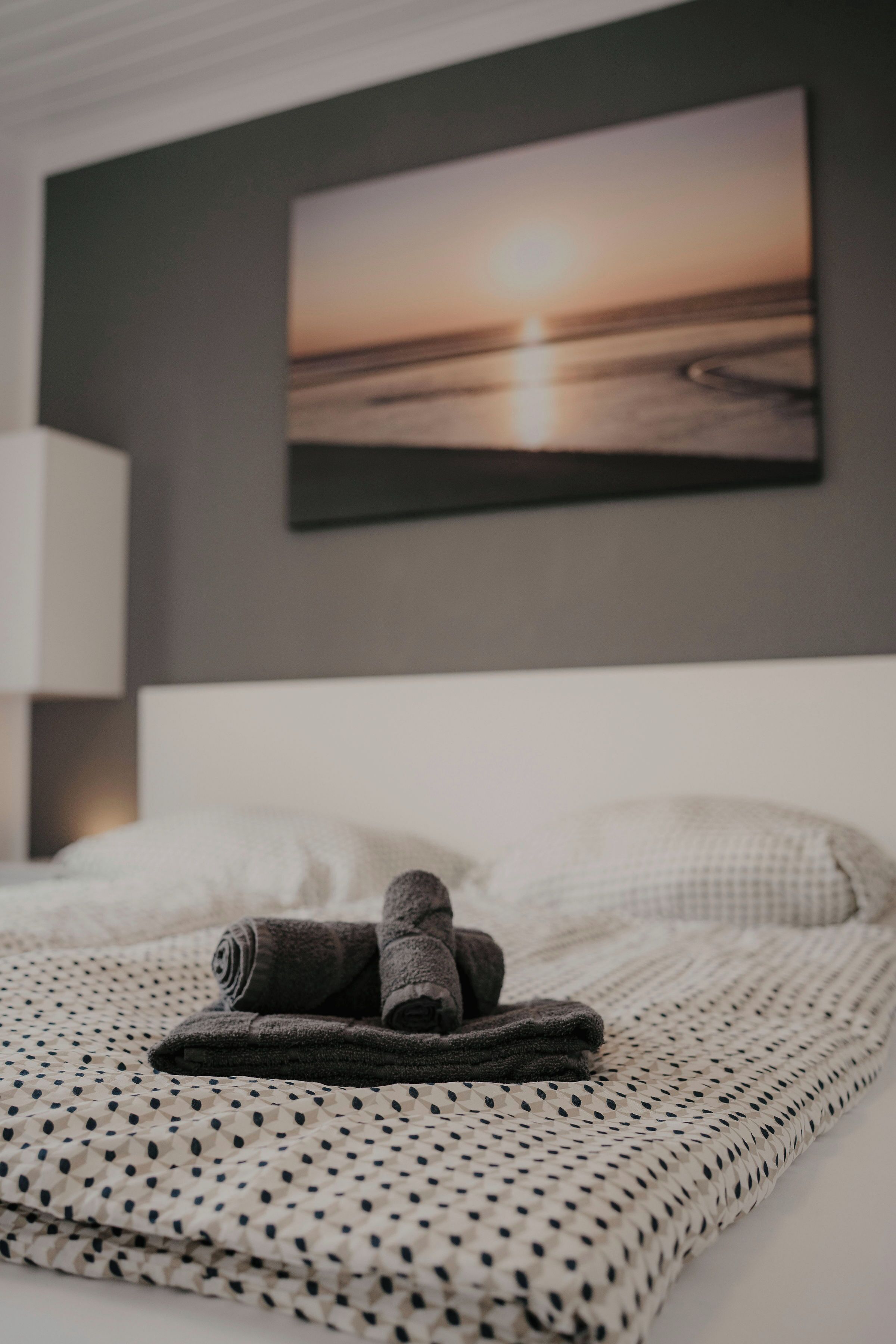 A neatly made bed with a patterned blanket, a rolled-up towel, and a painting of a sunset on the wall.
