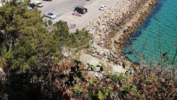 Un porto turistico con numerose imbarcazioni ormeggiate, un'area di parcheggio con auto e un litorale roccioso con acque cristalline blu.