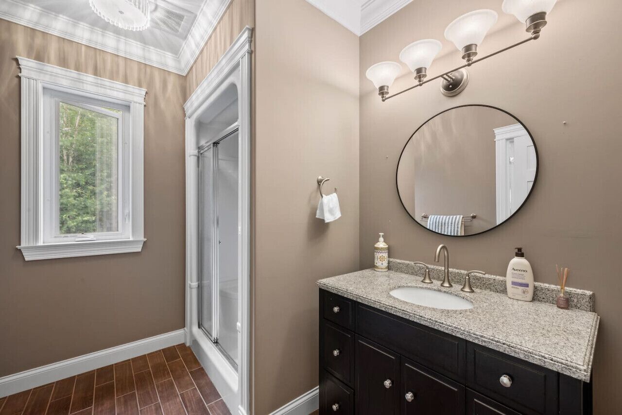 A bathroom with a large window, a shower, a sink with a vanity, and a mirror.