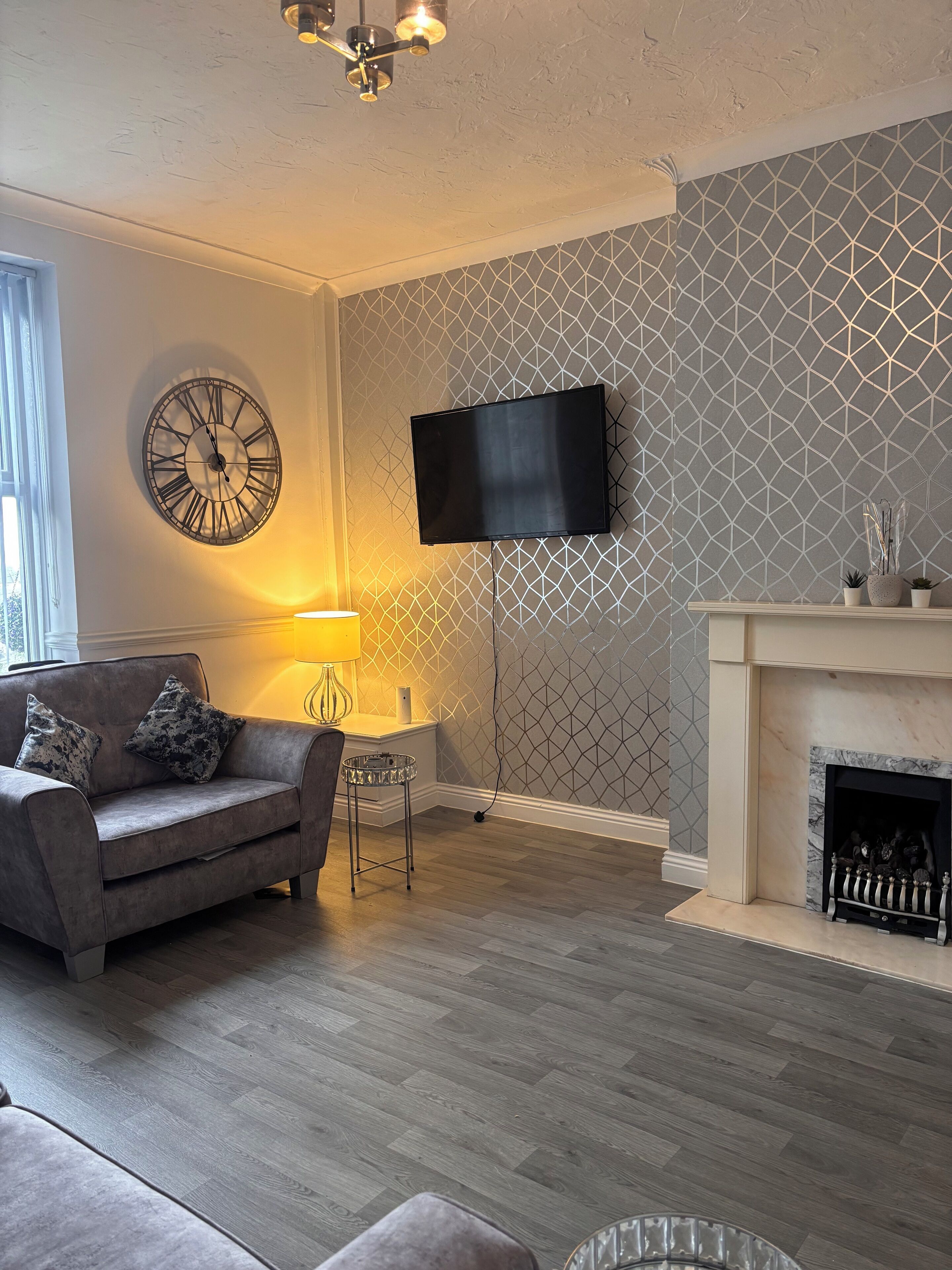A living room with a grey patterned wall, a mounted TV, a fireplace, a sofa with cushions, and a round wall clock.