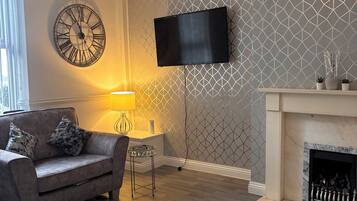 A living room with a grey patterned wall, a mounted TV, a fireplace, a sofa with cushions, and a round wall clock.