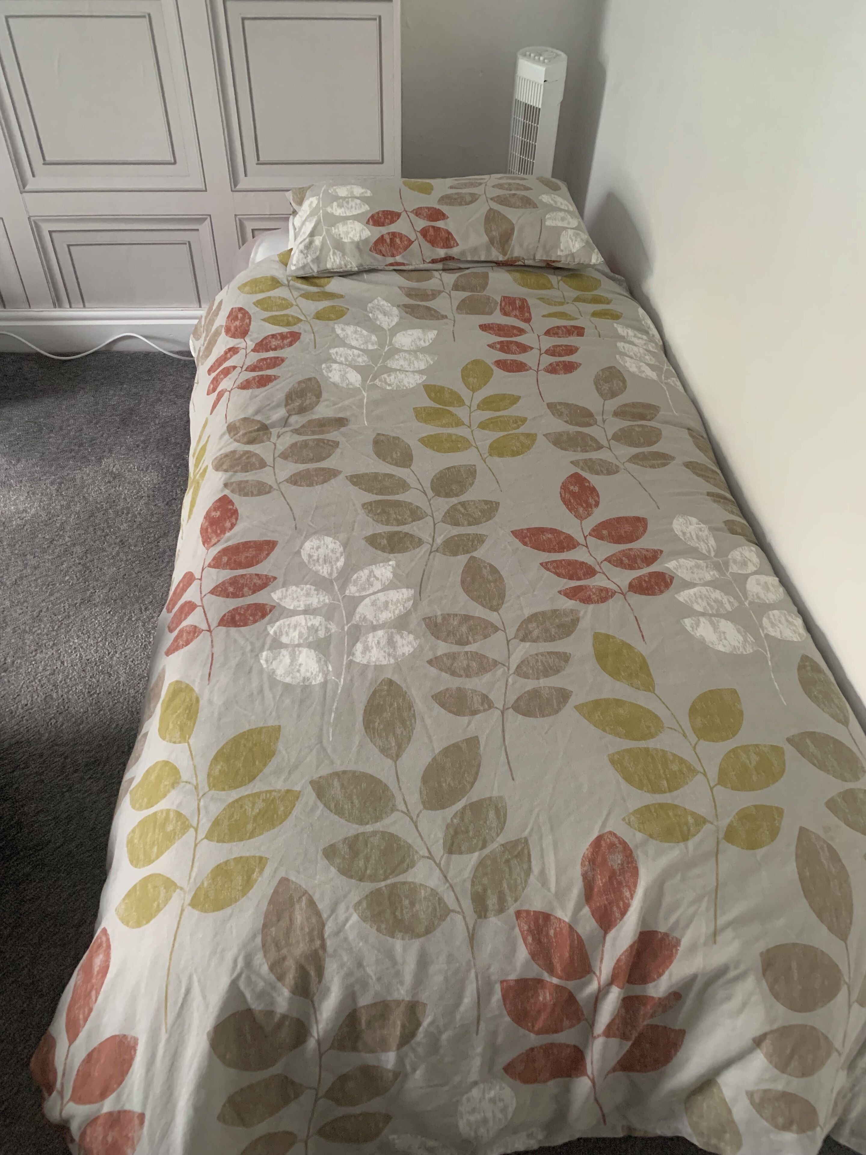 Een bed met een sprei en kussen met bladpatroon in een kamer met een witte garderobe en een witte radiator.
