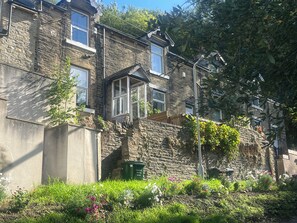 Une maison en pierre avec un jardin devant, un lampadaire et une poubelle verte portant l’inscription « Déchets ménagers ».