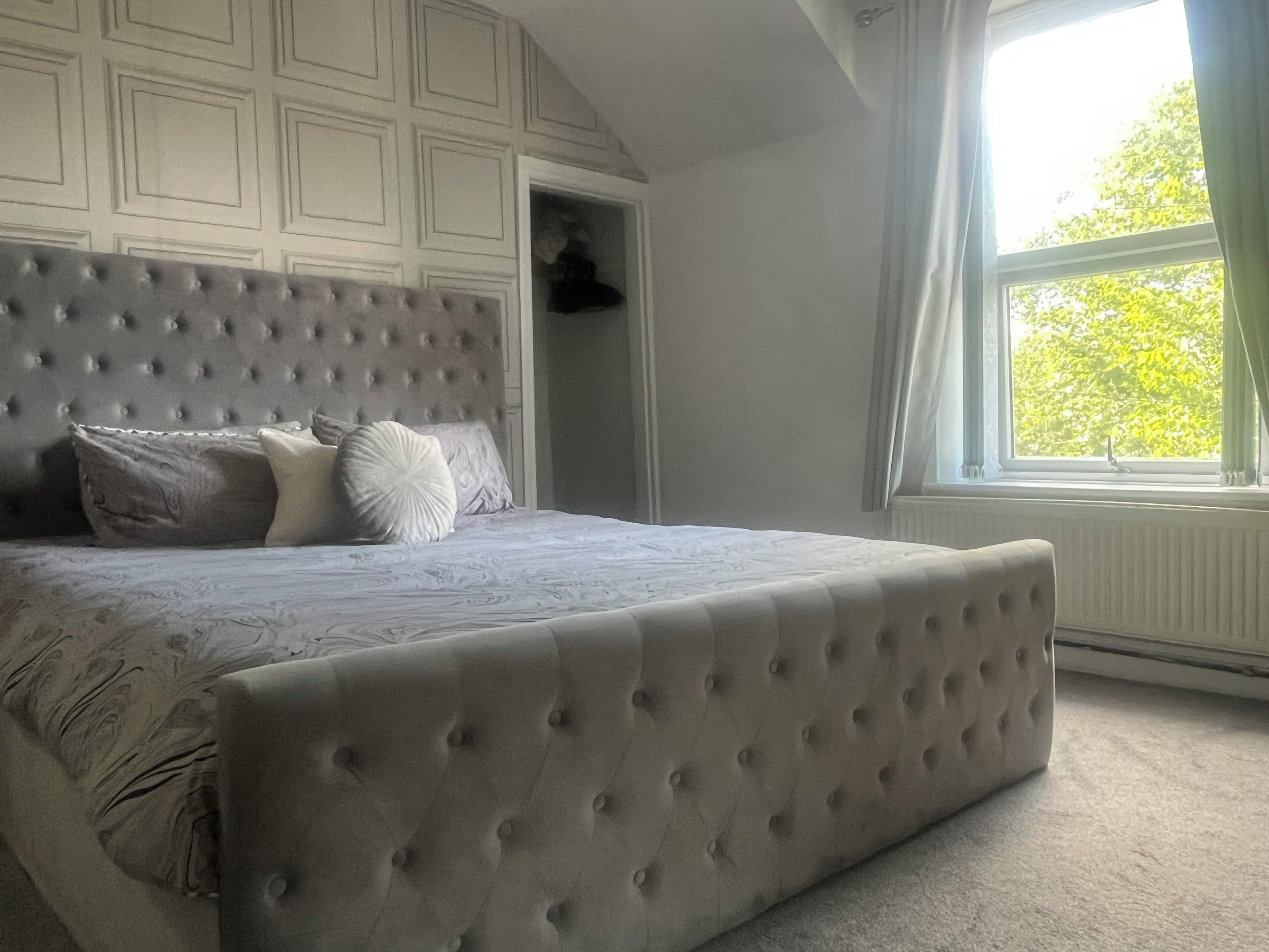 A bedroom with a tufted headboard, a bed with patterned bedding, a window with curtains, and a radiator.