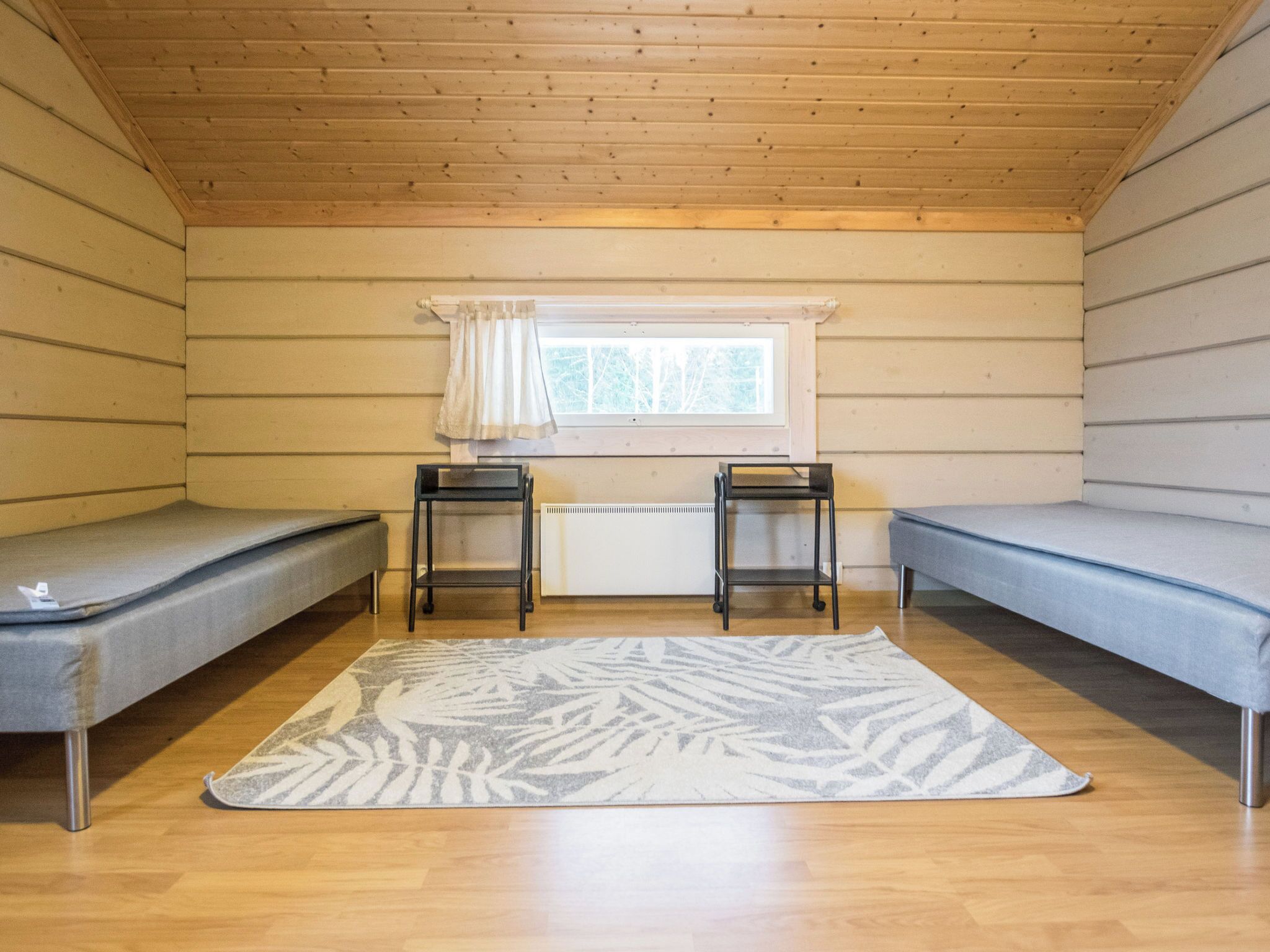 A small, single-room cabin with two bunk beds, a window with curtains, a radiator, and a patterned rug.