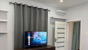 Quarto moderno com cómoda de madeira escura, uma televisão exibindo um filme, ventoinha no teto e cama com manta verde.