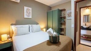 Ein Hotelzimmer mit Bett, Nachttischlampen, einem grünen Kleiderschrank, einem Badezimmer mit Waschbecken und Spiegel sowie Kunstwerken an den Wänden.