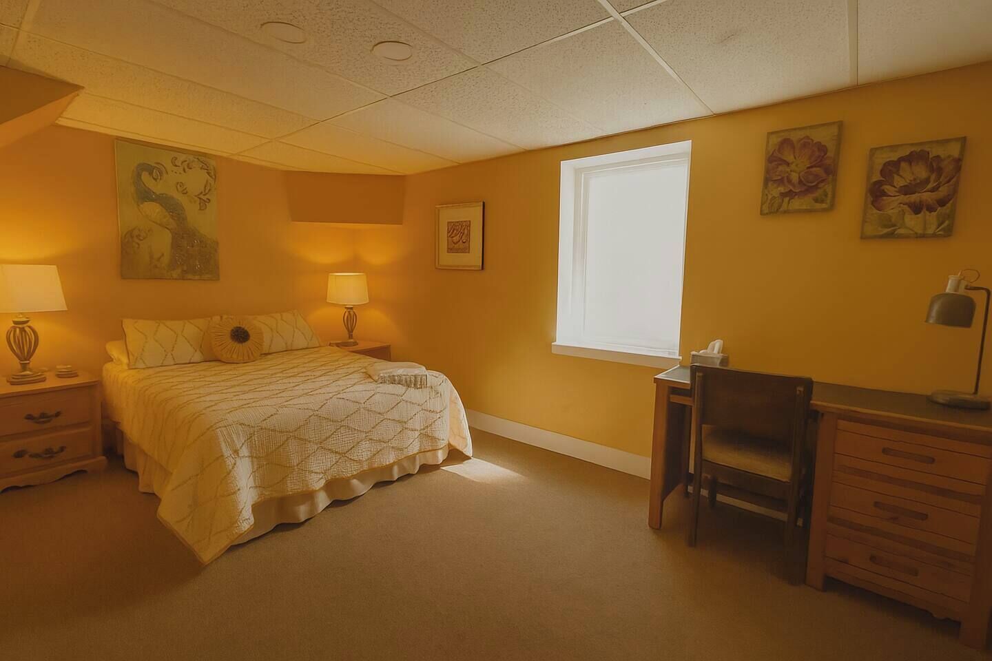 Une chambre à coucher avec un lit, des tables de chevet, un bureau et une chaise. Il y a deux fenêtres et des œuvres d’art accrochées au mur.