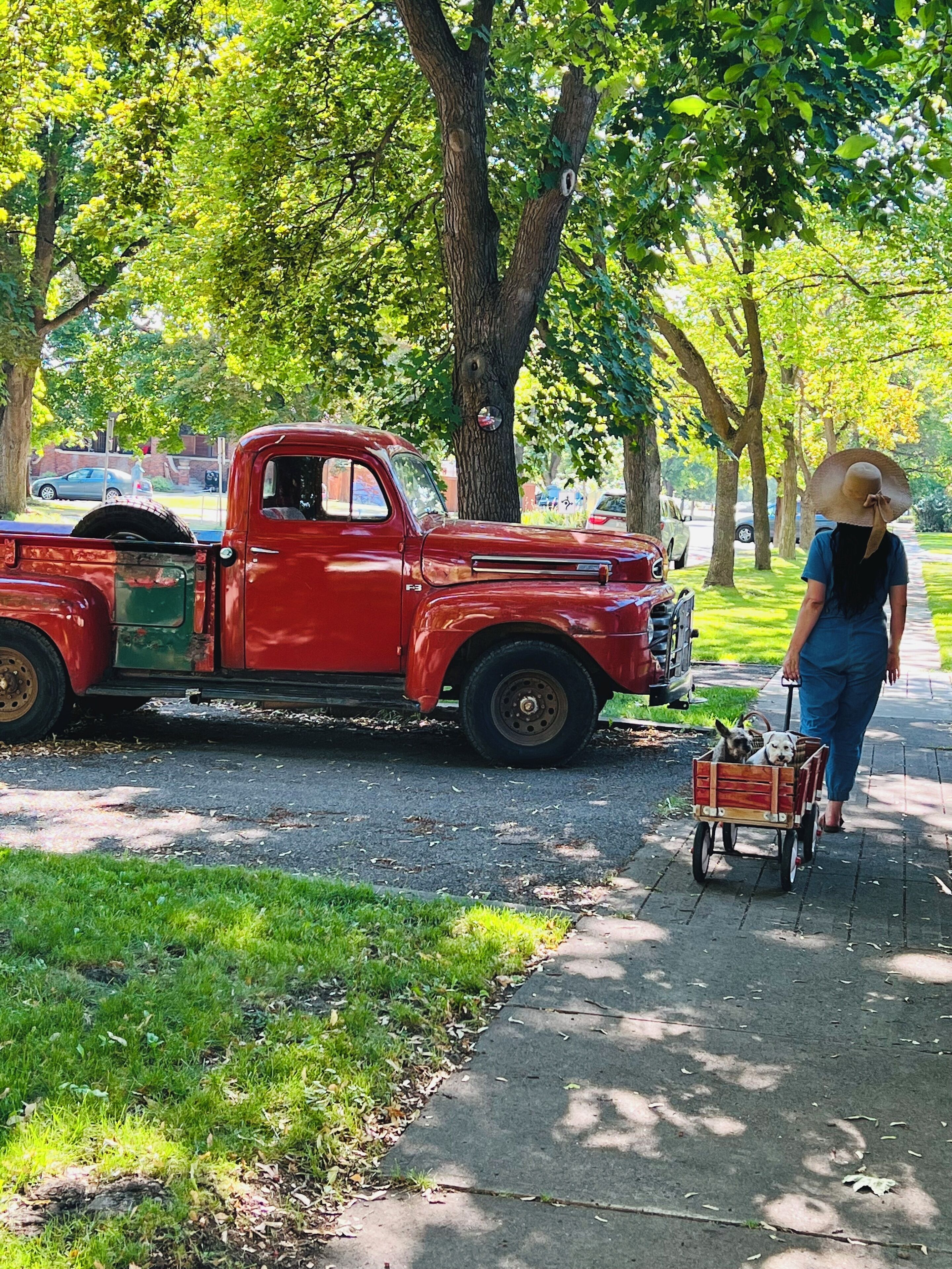 A red vintage truck parked on a street with a woman pushing a dog cart.