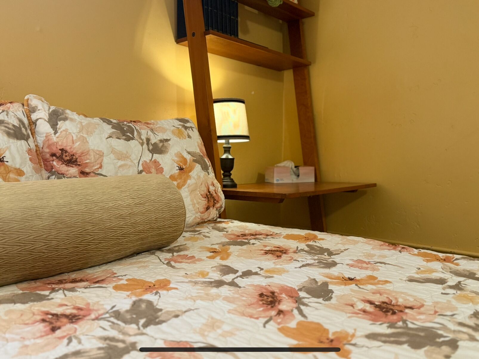 Een bed met een bloemenprint sprei en een geweven kussen, een nachtkastje met een lamp, en een spiegel aan de muur.