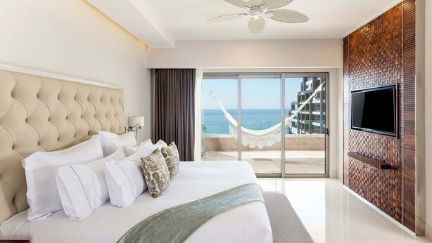 A modern hotel room with a large bed, a television, and a view of the ocean through a glass door. - Spacious 1‑bedroom suite, complete with premium 5‑star, resort‑style amenities. (Cabo San Lucas)
