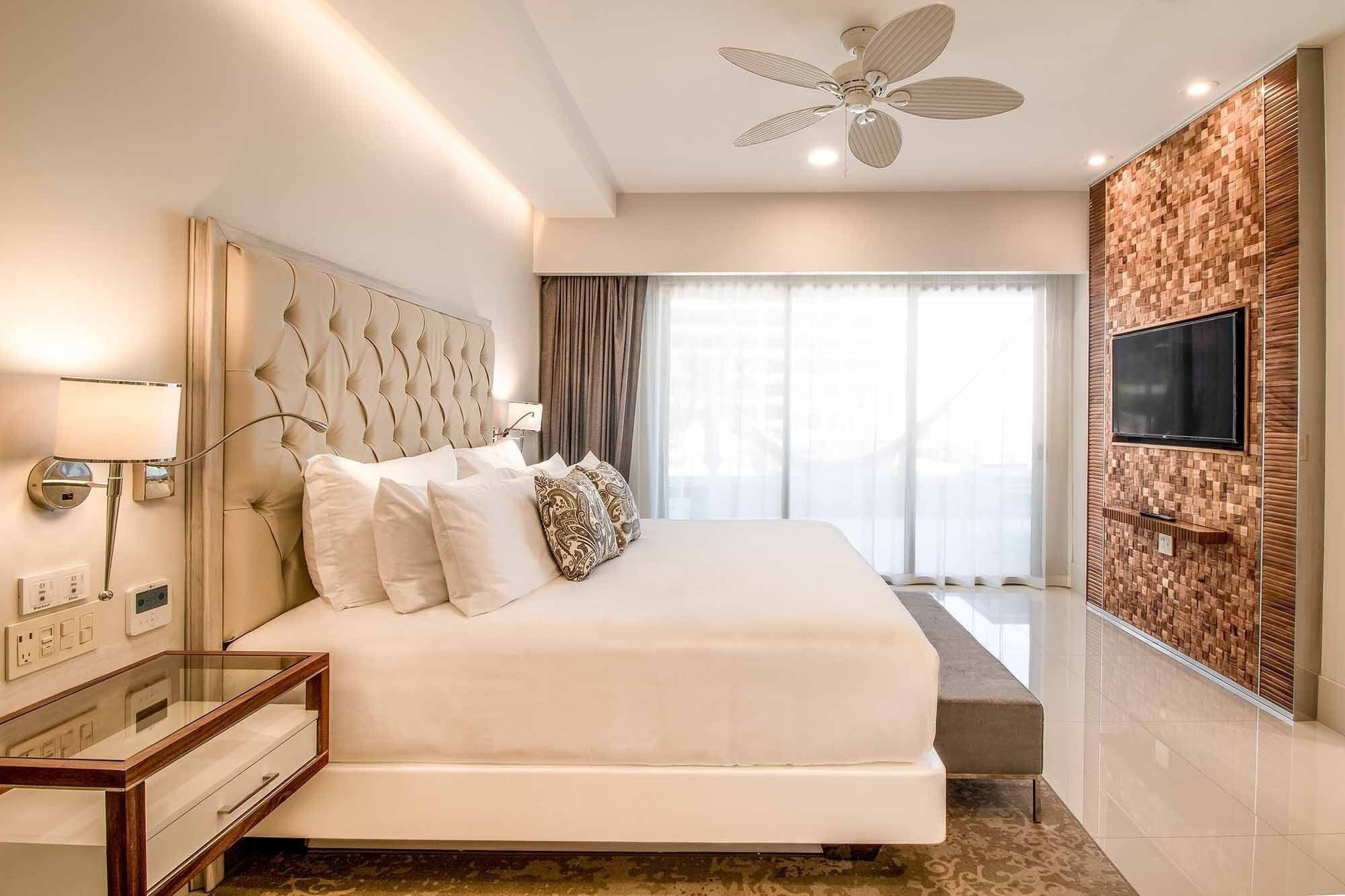 A modern hotel room with a large bed, a ceiling fan, a TV mounted on the wall, and a window with curtains.