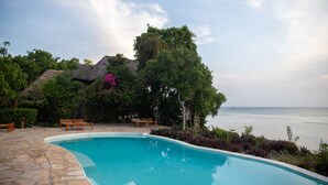 A swimming pool with a stone deck, wooden lounge chairs, and a view of the sea. - Veridiana Hotel (Kizimkazi)