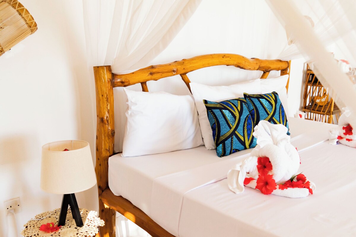 Un lit avec une tête de lit en bois, une literie blanche et un baldaquin. Il y a une table de chevet avec une lampe et un objet décoratif.