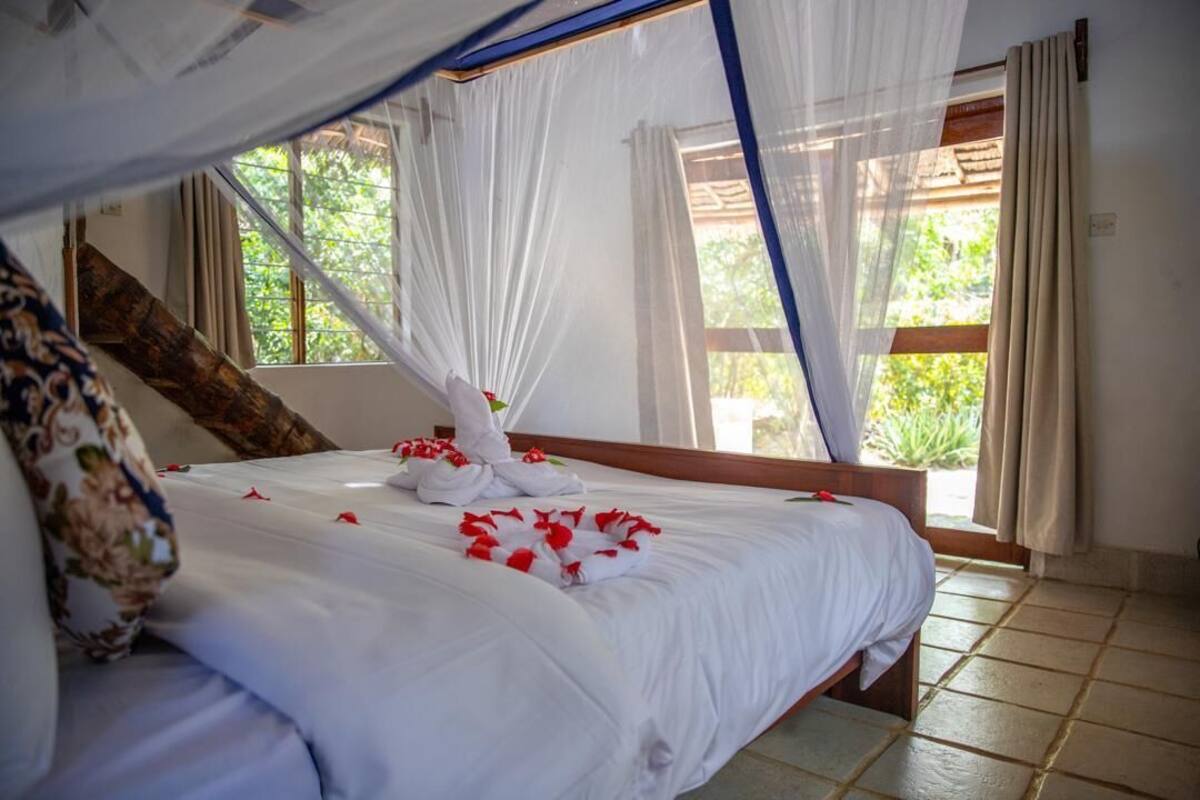 Un lit bien fait, avec du linge de lit blanc et des pétales de roses rouges, une tête de lit en bois et une fenêtre donnant sur un paysage verdoyant.