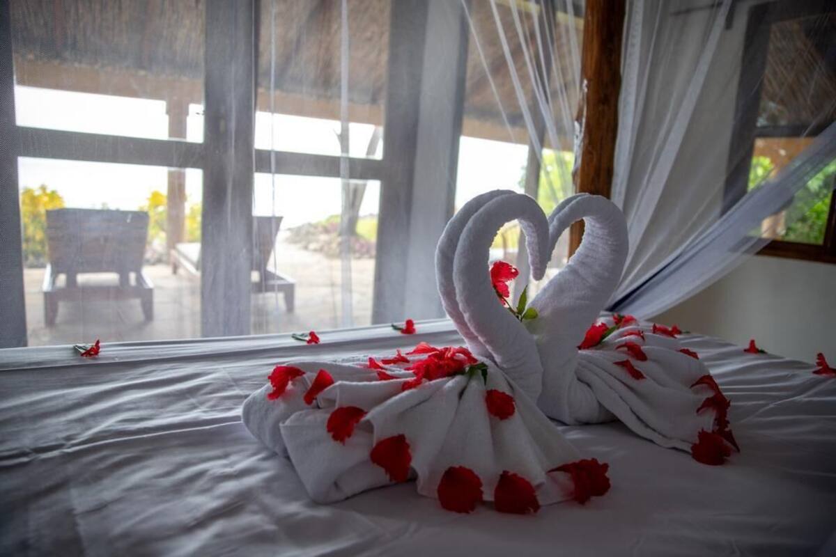 Un lit agrémenté d’une décoration en forme de cœur faite avec des serviettes et des pétales de rose, une fenêtre donnant sur des meubles d’extérieur et des rideaux légers.