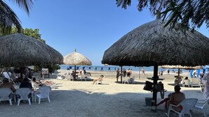 Una playa con sombrillas de paja, sillas de plástico blancas y gente relajándose.