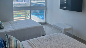 Habitación de hotel con dos camas, televisión y un amplio ventanal con vistas a edificios y montañas.