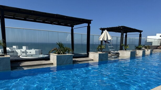 Una piscina infinita en la azotea con barandilla de cristal, sillas de descanso y una zona de estar cubierta.