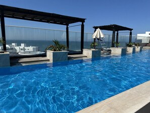 Une piscine à débordement située sur le toit, avec une balustrade en verre, des chaises longues et un espace salon couvert.