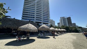 Una playa con sombrillas de paja y sillones de descanso, un alto edificio de hotel y una grúa de construcción al fondo.